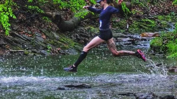 A runner leaps across a stream on a wooded trail, with water splashing around their feet.