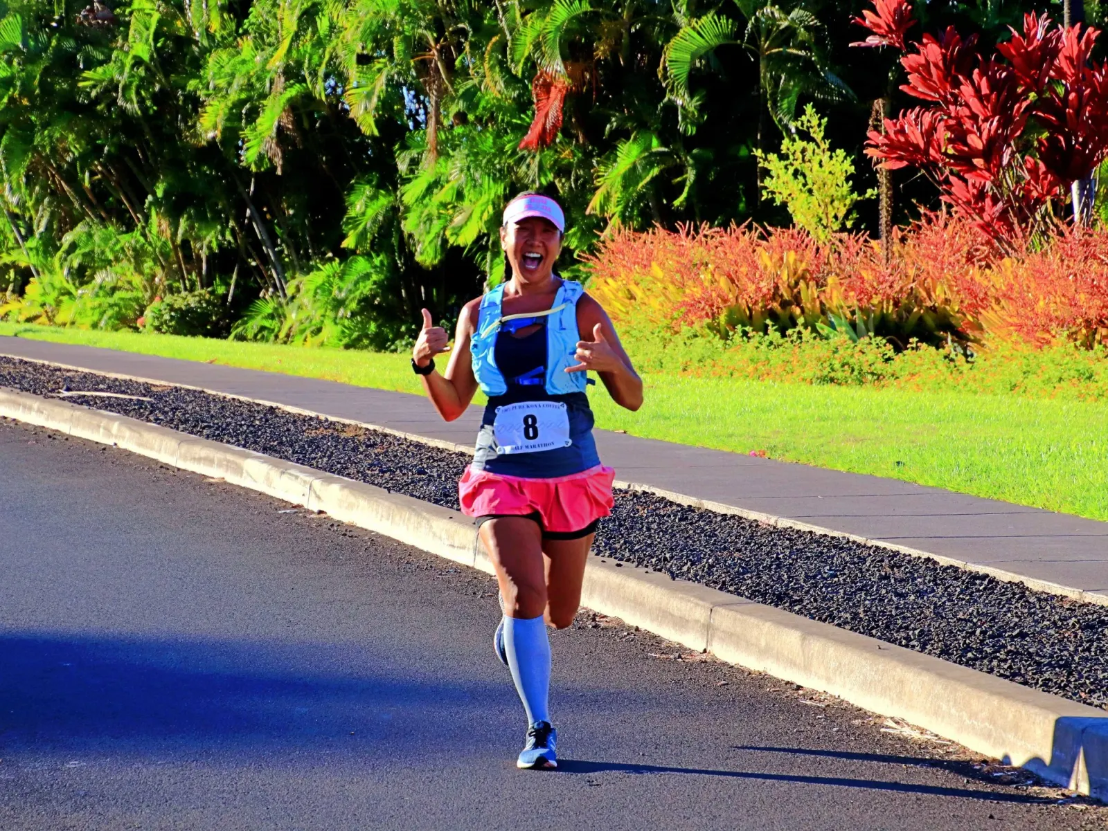 A female runner with a bib number 8 smiles and gives a thumbs up while running on a road lined with tropical foliage.