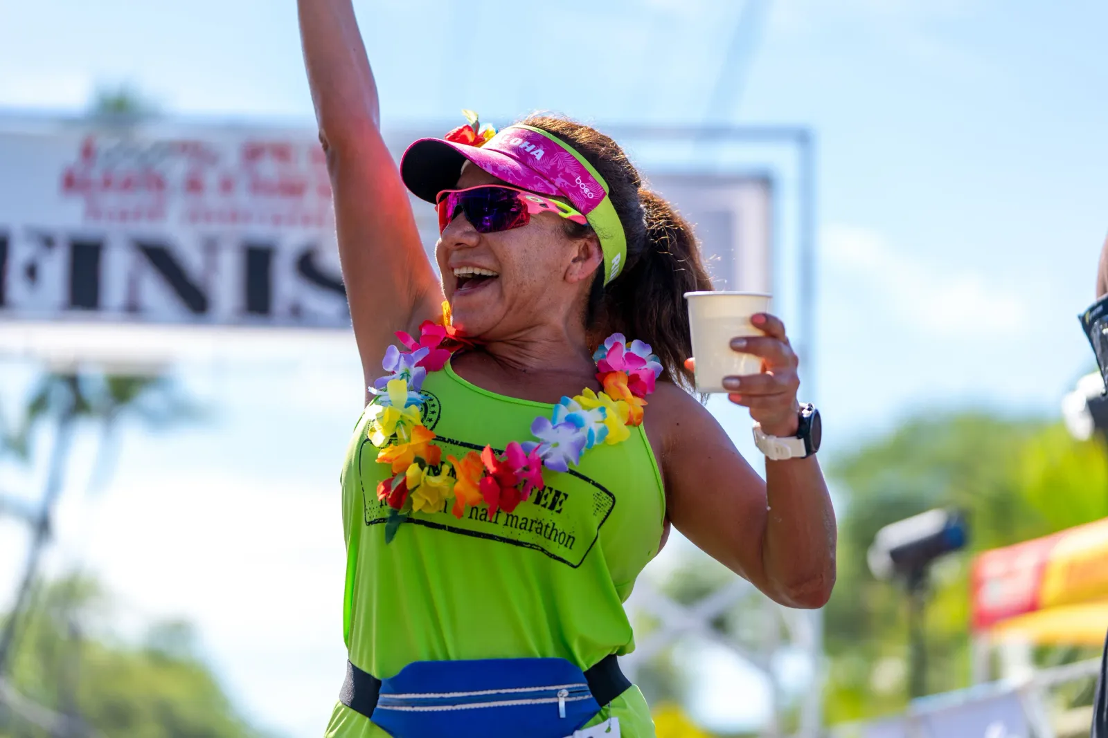 A smiling woman with a lei around her neck and a visor on her head raises her arm in celebration as she crosses the finish line of a race, holding a cup.