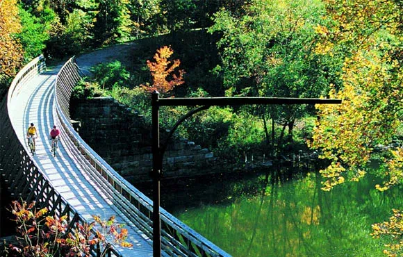 Two cyclists on a wooden bridge over a green body of water, surrounded by trees in autumn colors.