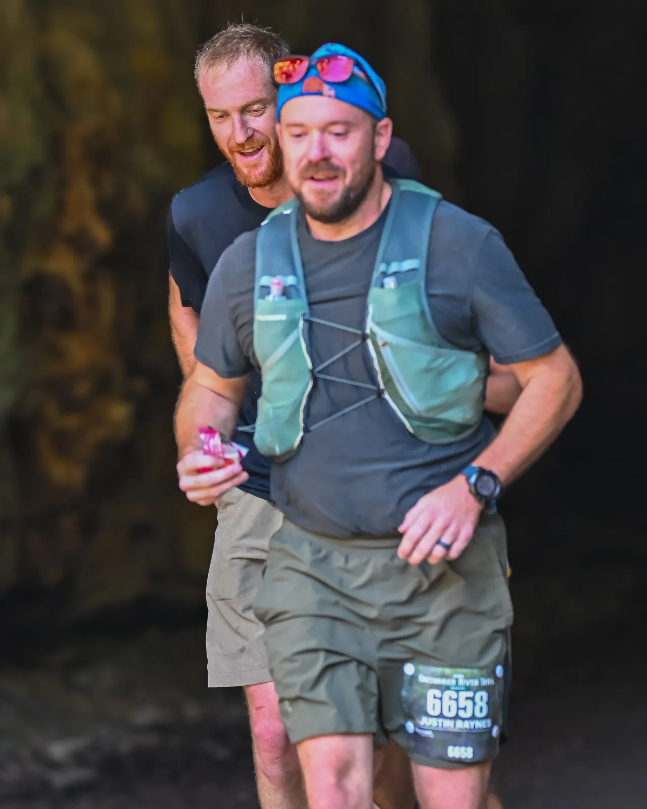 Two male runners on a trail, one in front wearing a hydration vest and bib number 6658, the other behind him smiling.