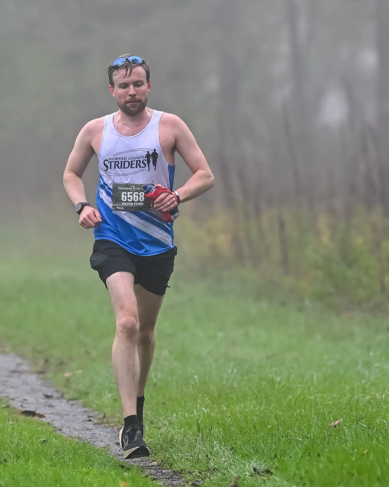A male runner wearing a white and blue tank top with the number 6568 and sunglasses on his head runs on a path next to a grassy area.
