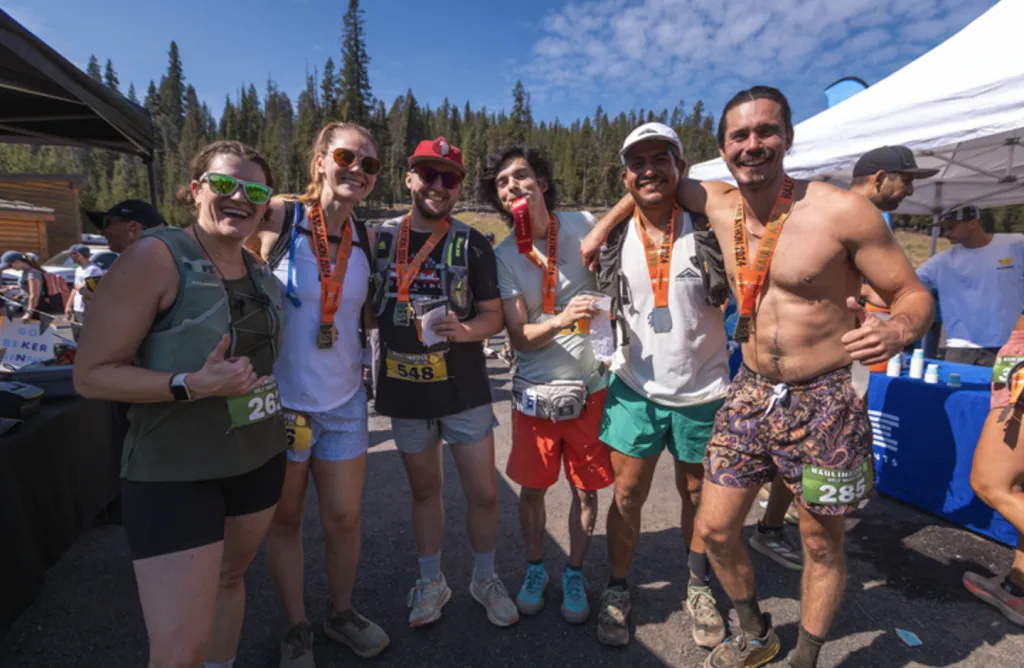 A group of six smiling runners, some wearing medals, pose for a photo after a race, with trees and tents in the background.