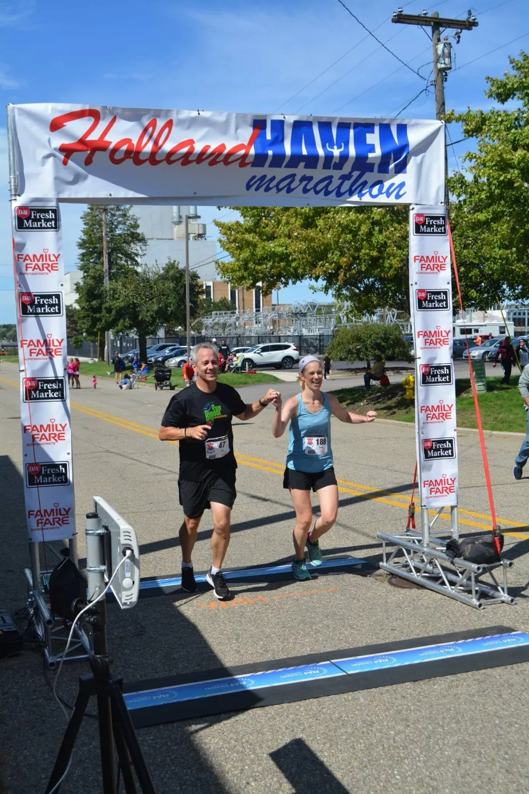 A male and female runner, holding hands, cross the finish line of the Holland Haven Marathon under a large banner.