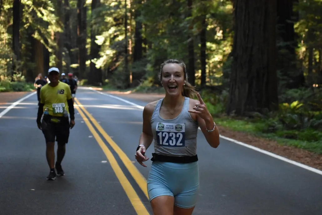 A female runner smiles and makes a rock-on hand gesture while running on a road through a redwood forest.