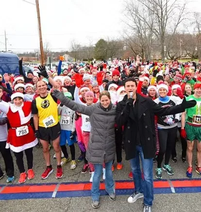 A large crowd of runners, many wearing Santa hats, are gathered at the start line of a race, with two people in the foreground holding microphones and waving.