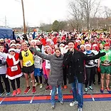 A large group of runners wearing Santa hats and festive attire are gathered at the start line of a race, with two people in the foreground addressing the crowd.