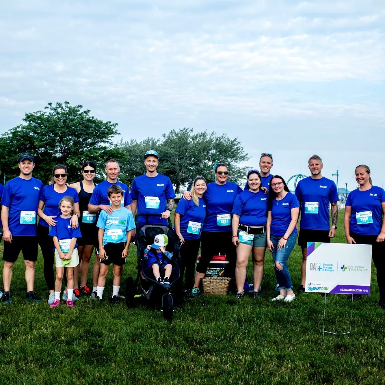 A group of runners in blue shirts, some with race bibs, pose for a photo on a grassy field with trees in the background.