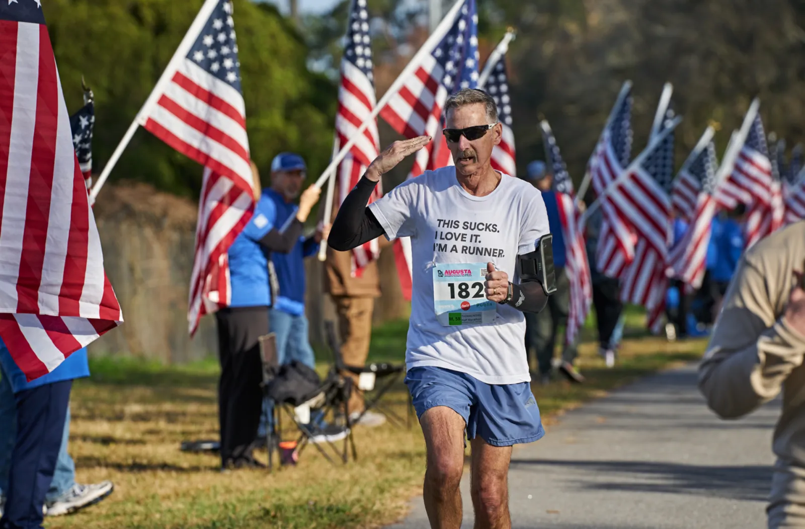 A male runner salutes while running past American flags, wearing a t-shirt that says 'THIS SUCKS. I LOVE IT. I'M A RUNNER.'