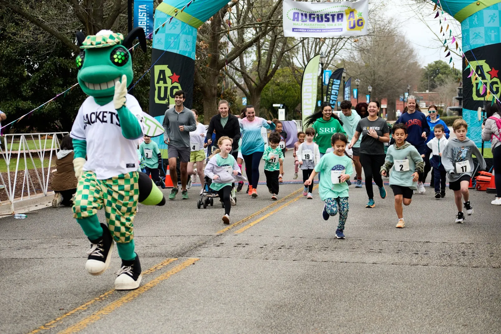A mascot leads a group of runners, including children, across the start line of the Augusta Half Marathon.