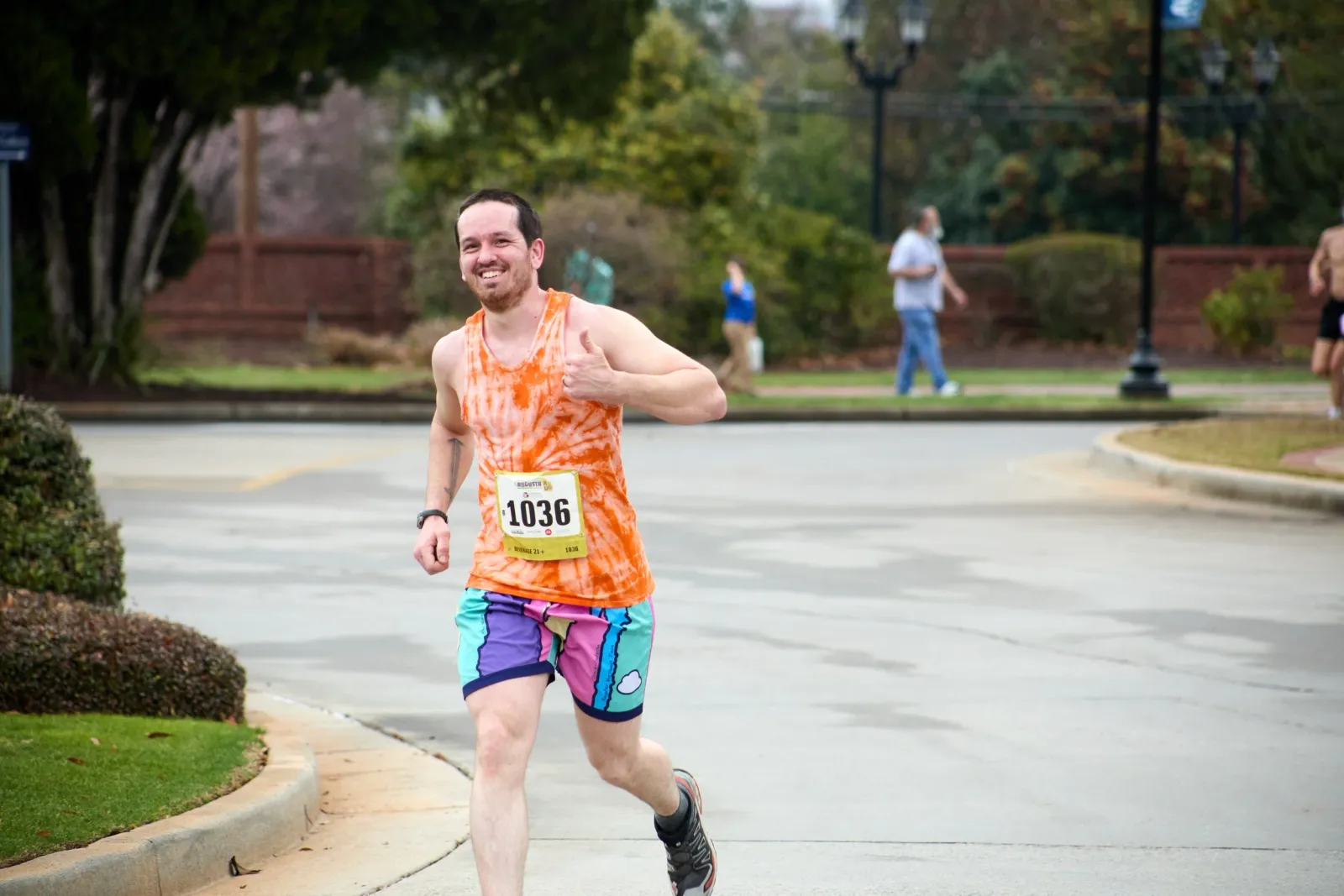 A male runner in an orange tie-dye tank top and colorful shorts smiles and gives a thumbs up while running on a road during a race.