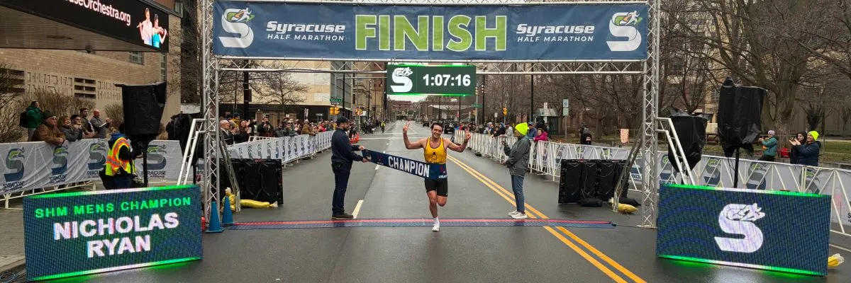 A male runner, Nicholas Ryan, wearing a yellow tank top and black shorts, crosses the finish line of the Syracuse Half Marathon, holding up peace signs with both hands. He is breaking through a banner that reads "CHAMPION". A digital clock above shows "1:07:16". Spectators are visible on either side of the course.