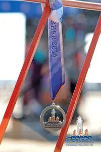 A finisher's medal for the 3 Beach Minimum Half Marathon hangs on a red railing, with the blurred finish line and crowd in the background.