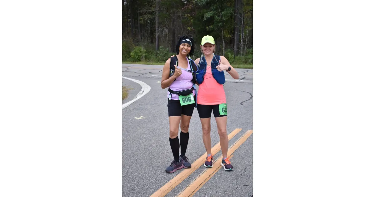 Two female runners posing for a photo on a road course, both smiling and giving thumbs up.