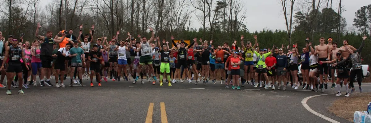 A large group of runners jump in the air with their arms raised, celebrating at the start or finish line of a road race.