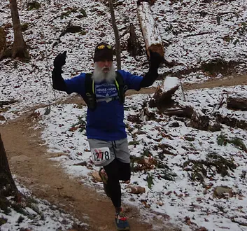 A runner with a long white beard and a blue t-shirt runs on a snowy trail with his arms raised in excitement.