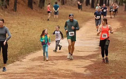 Runners of various ages, including children, participating in a race on a dirt path through a forest.
