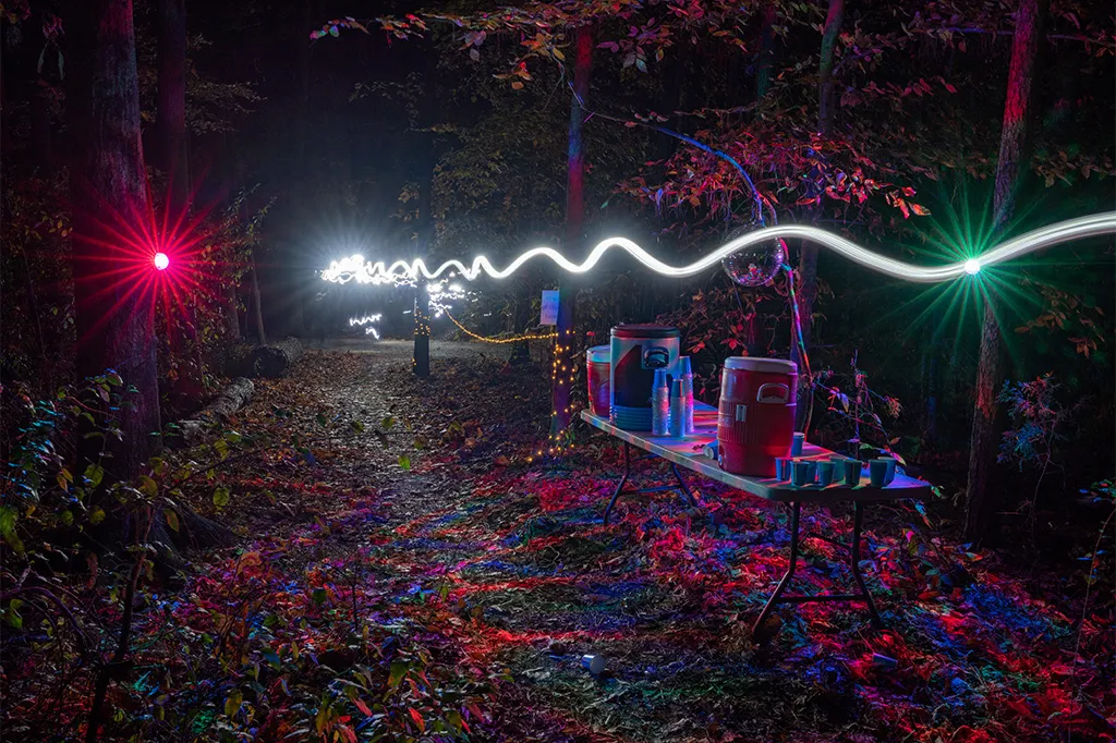 A forest trail at night illuminated by colorful lights and a wavy light streak, with a table set up with coolers and cups.