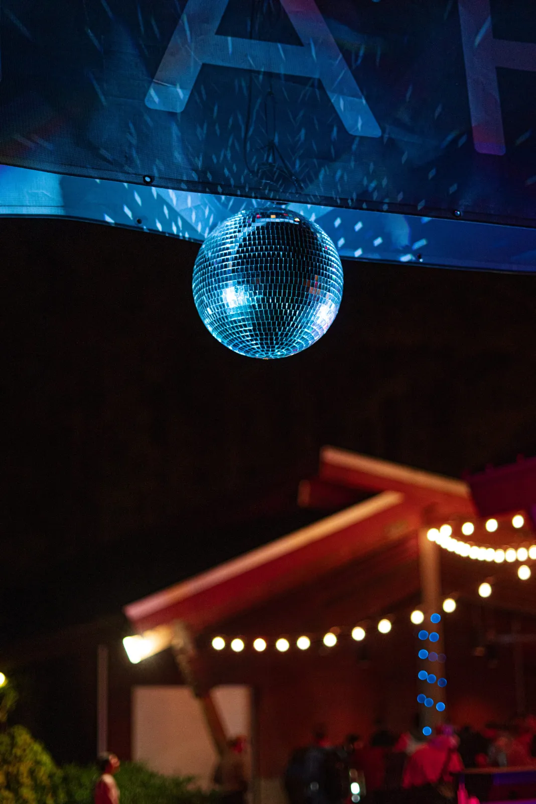 A disco ball hangs in the air, illuminated by blue lights, with a blurred background of string lights and people at a night event.