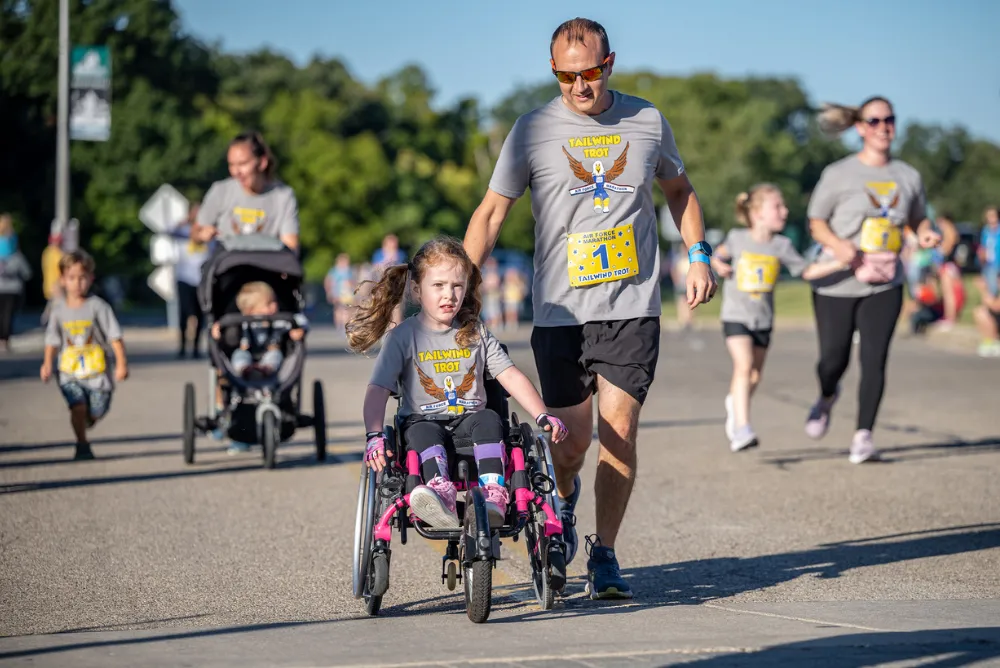 A father runs alongside his daughter in a wheelchair during a race, both wearing matching t-shirts.