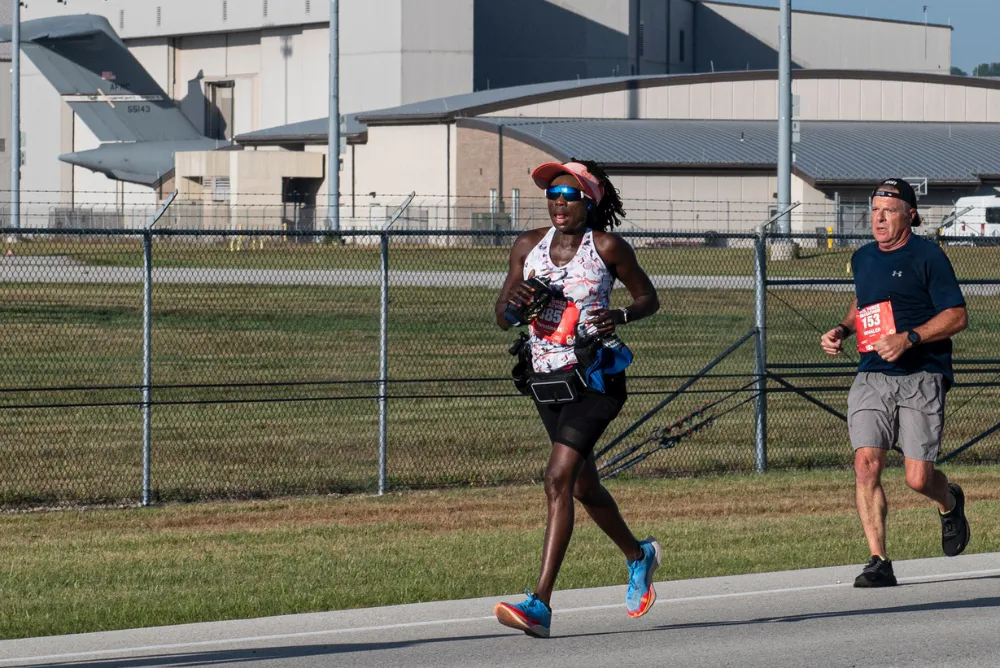Two runners, a woman in the foreground and a man behind her, running on a road with an airplane and buildings in the background.