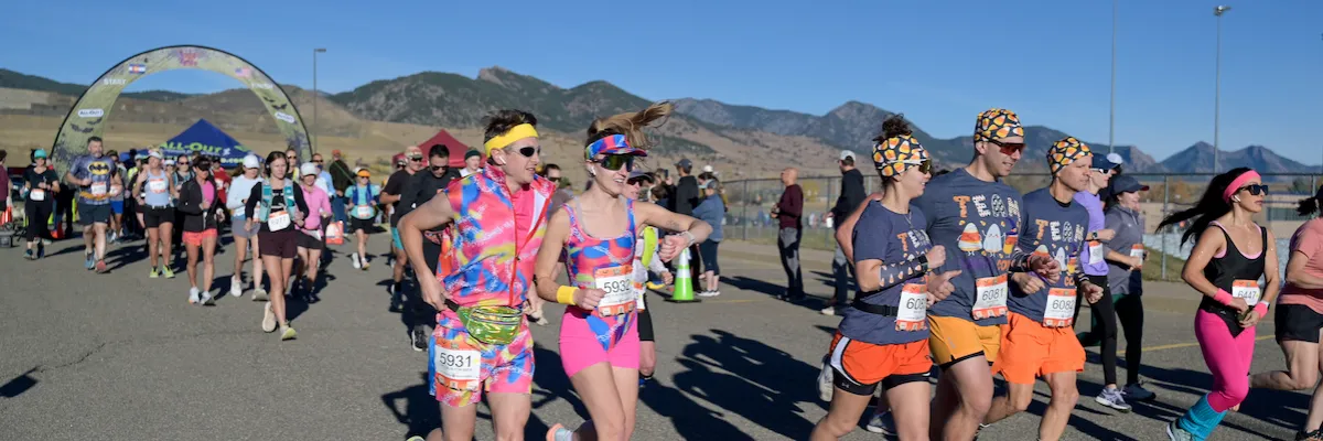 Runners in colorful costumes and athletic wear start a race under a festive archway, with mountains in the background.