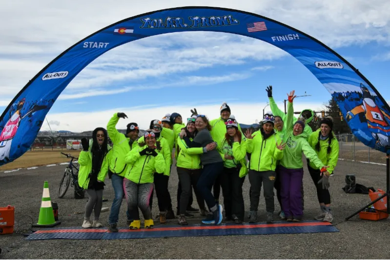 A group of people in bright yellow jackets and penguin hats pose and cheer under a blue race arch that says "Polar Prowl" with "START" and "FINISH" marked.