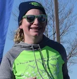 A runner wearing a beanie, sunglasses, and a green and grey hoodie with a mountain graphic and text that says 'If You're Not Going to Go All-Out'.