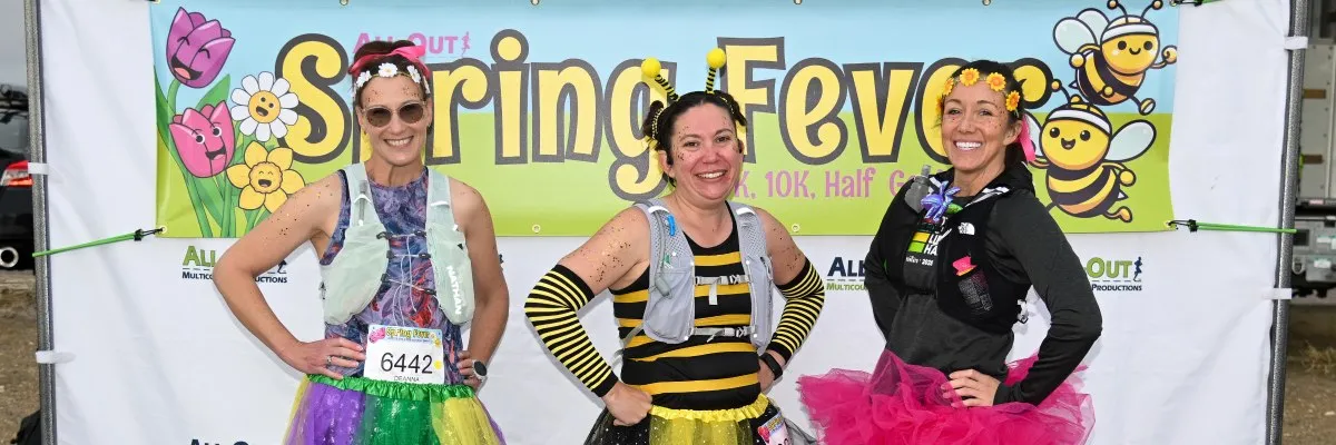 Three women in colorful outfits and race bibs smiling in front of a 'Spring Fever' banner.