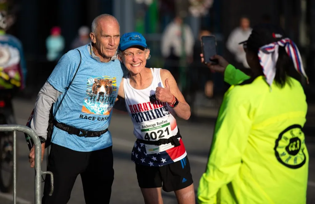 Two runners, a man and a woman, pose for a photo with the woman giving a thumbs up, with a third person taking their picture.