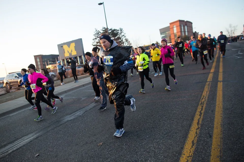 Runners participating in the Ann Arbor Marathon, with a runner in a black trash bag costume in the foreground and the University of Michigan 'M' logo visible in the background.
