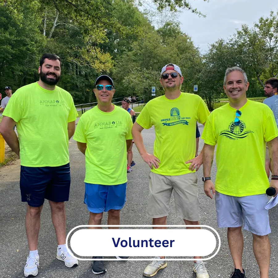 Four men in bright yellow t-shirts stand together, smiling, with a subtle 'Volunteer' overlay at the bottom.