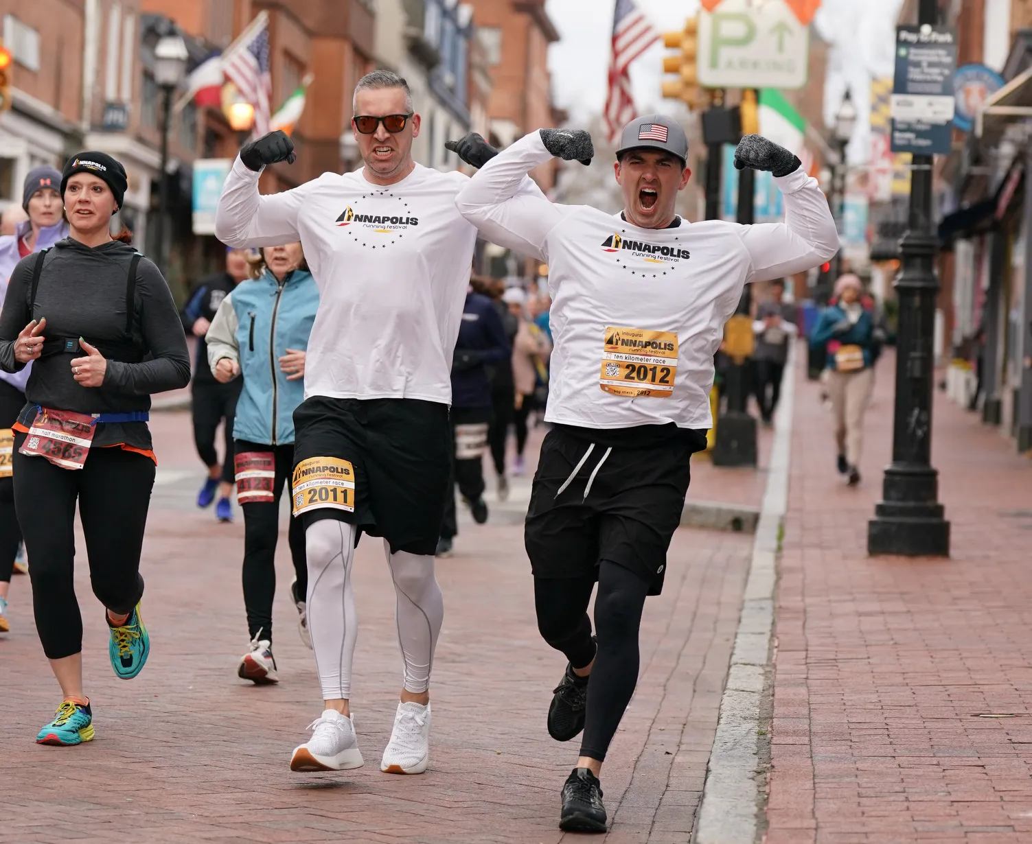 Two male runners celebrating with arms raised while running in a race, with other runners and spectators in the background on a brick street.
