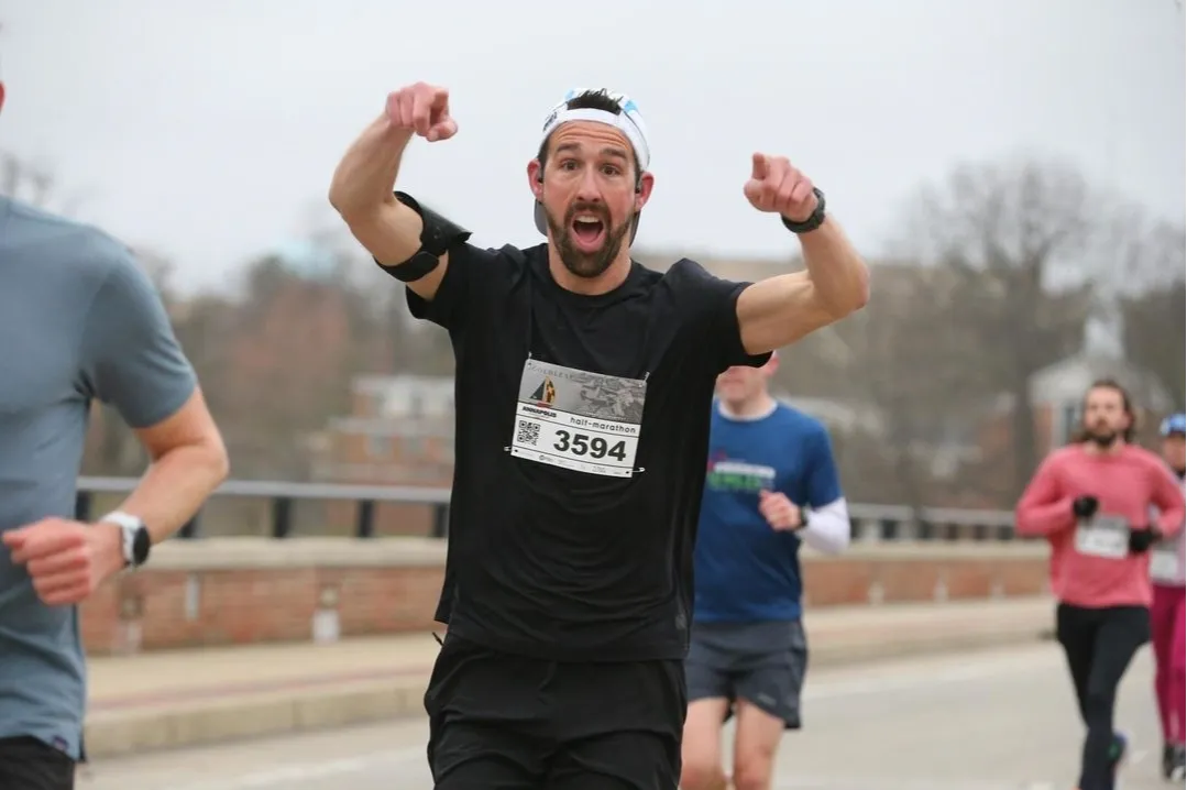 A male runner in a black t-shirt and white headband pumps his fists in the air and points forward with an excited expression, wearing bib number 3594 during a half-marathon.
