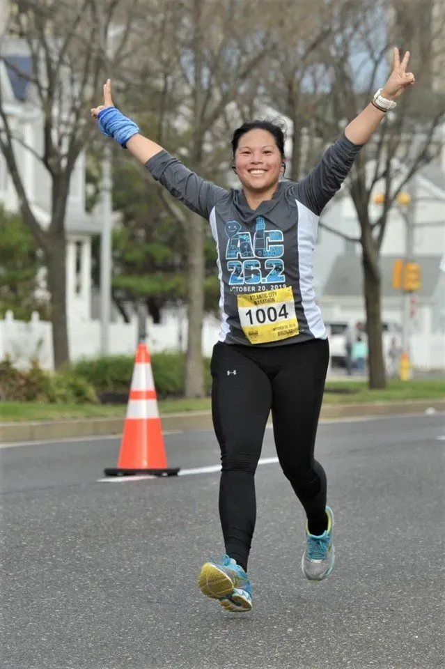 A female runner with arms raised in celebration, wearing bib number 1004 and a race t-shirt, smiles at the camera while running on a road.