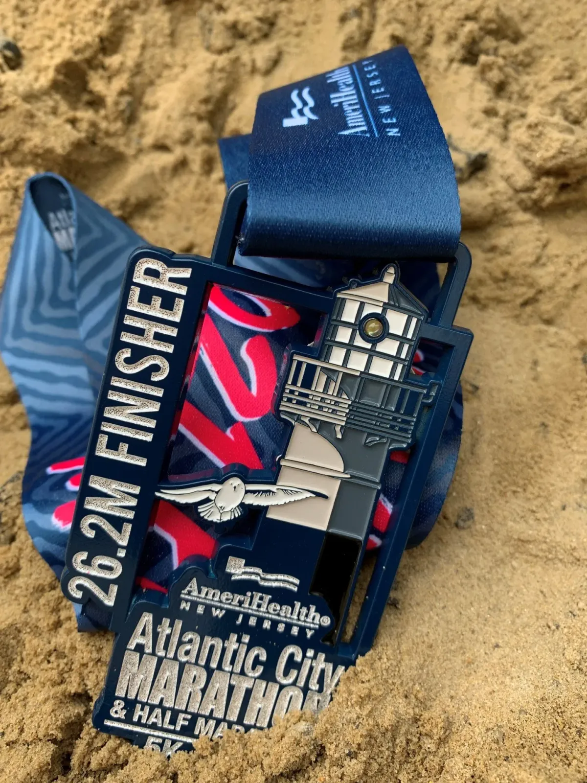 A close-up photo of the Atlantic City Marathon & Half Marathon medal lying on sand. The medal features a lighthouse design and the text '26.2M FINISHER'.