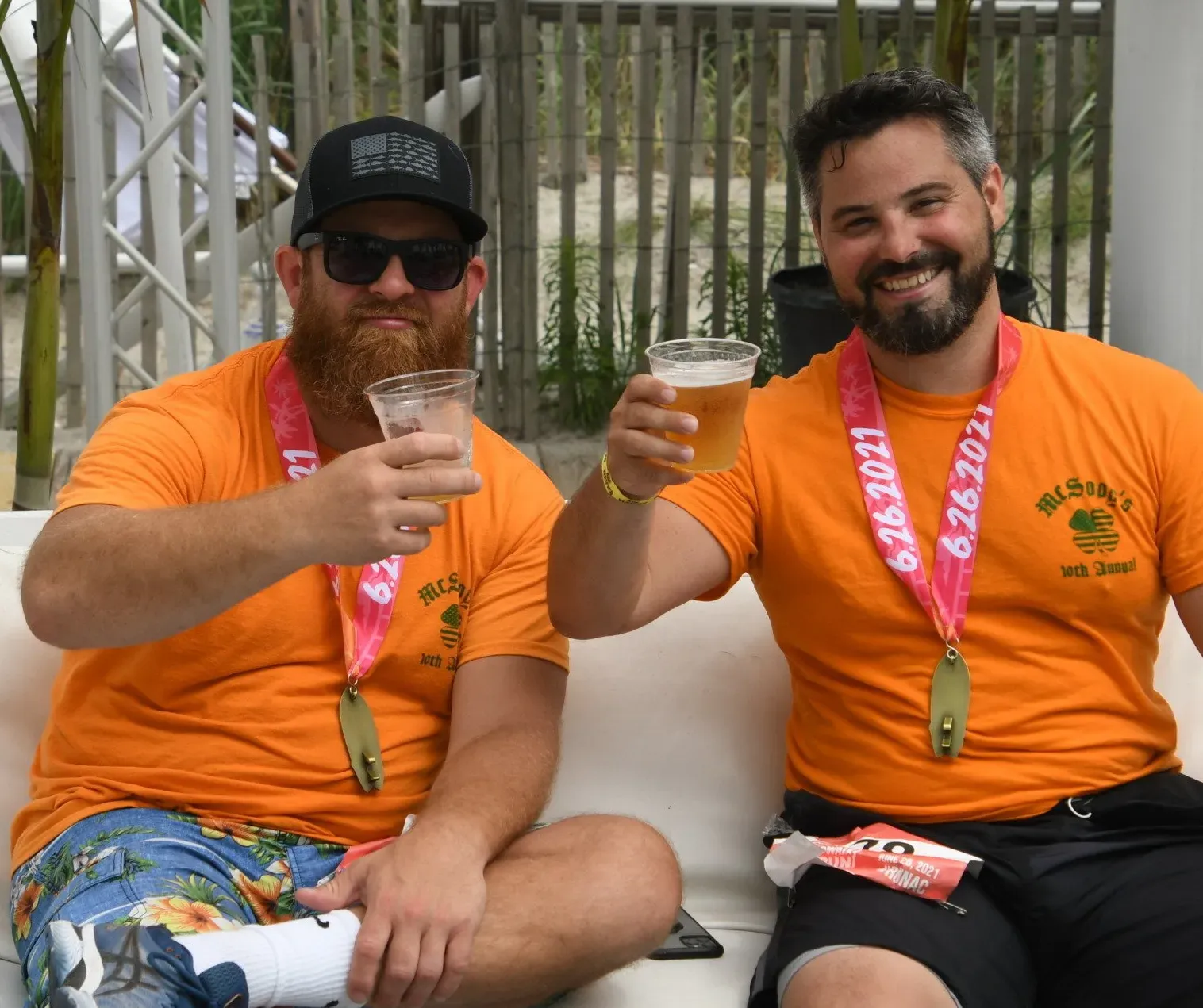 Two men wearing orange t-shirts and race medals are sitting and holding beers, smiling.