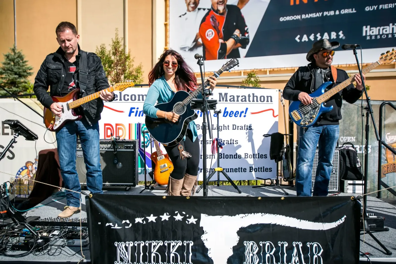 A band performs on a stage at a race event, with musicians playing guitars and a bass, and a banner in the foreground.