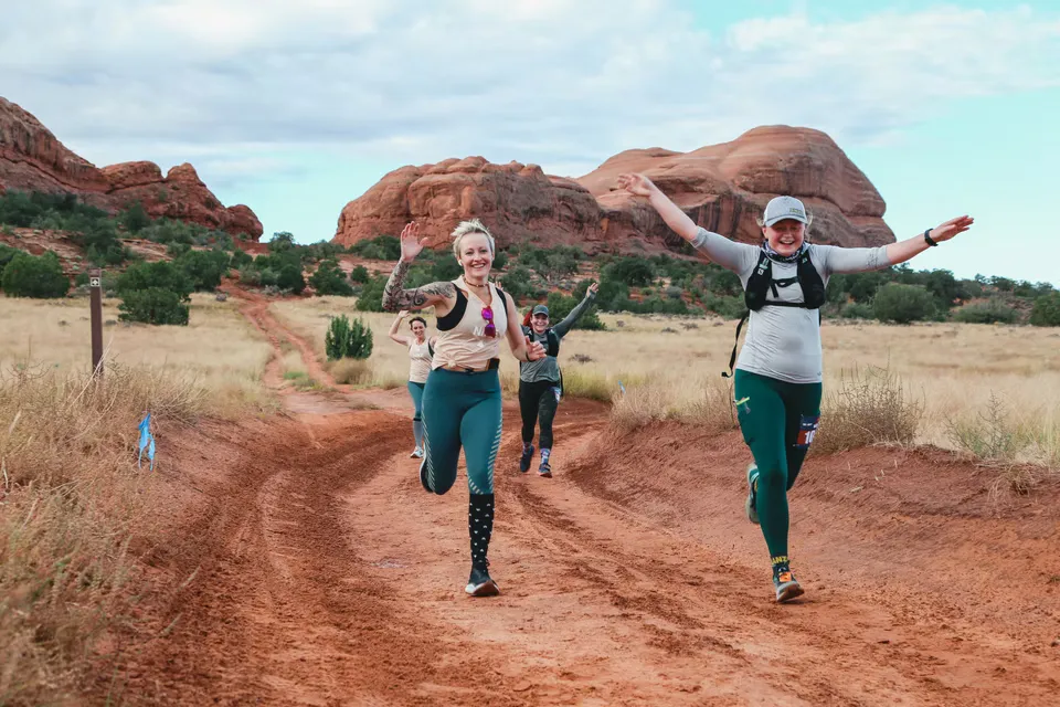 Runners smiling and waving as they run on a dirt trail with red rock formations in the background.