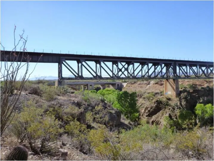 A large metal train bridge spans a dry, rocky canyon with desert vegetation in the foreground under a clear blue sky.