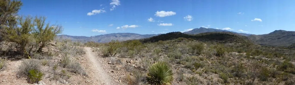 A panoramic view of a dusty trail winding through a desert landscape with mountains in the background under a blue sky with scattered clouds.