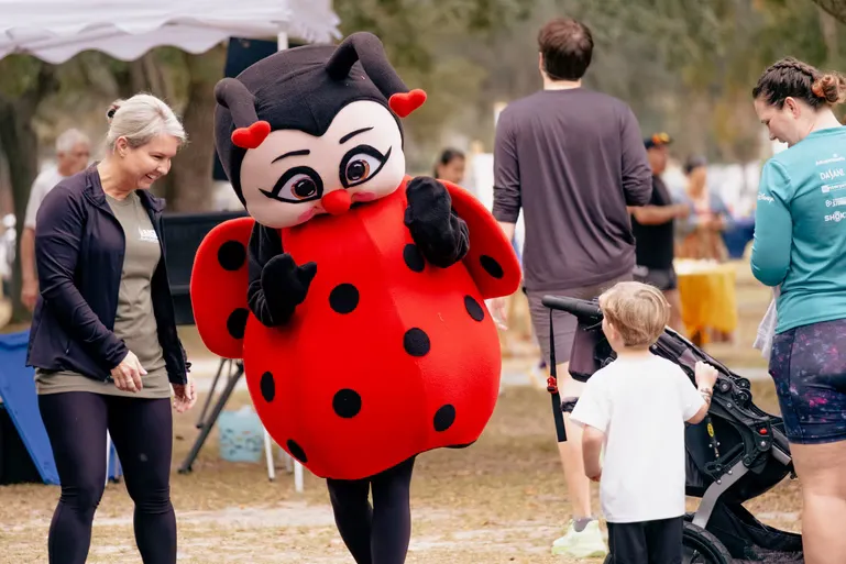 A person in a ladybug mascot costume interacts with a woman and a child at an outdoor event.