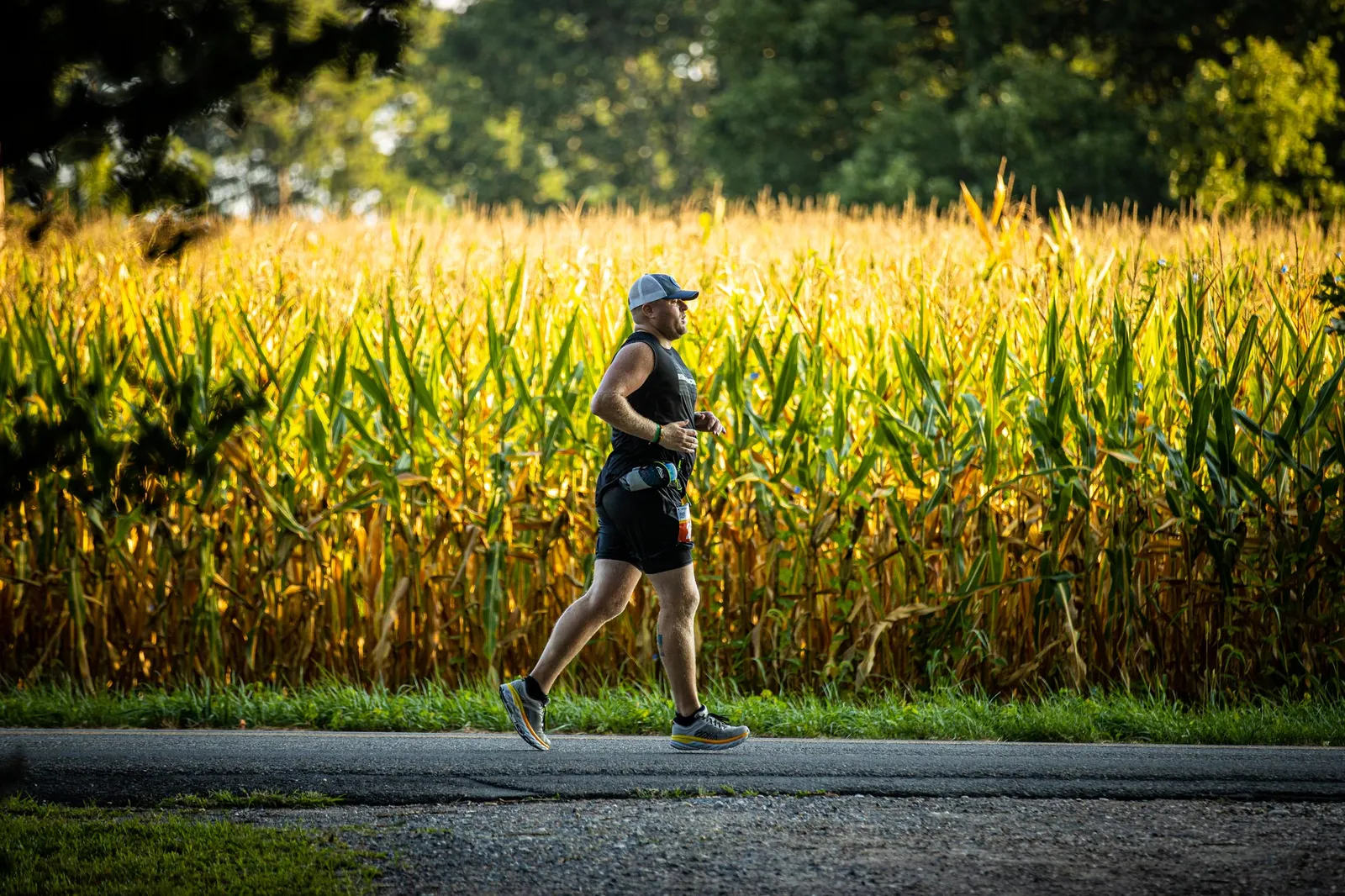 A male runner in a black tank top and shorts runs on a paved road next to a tall cornfield.