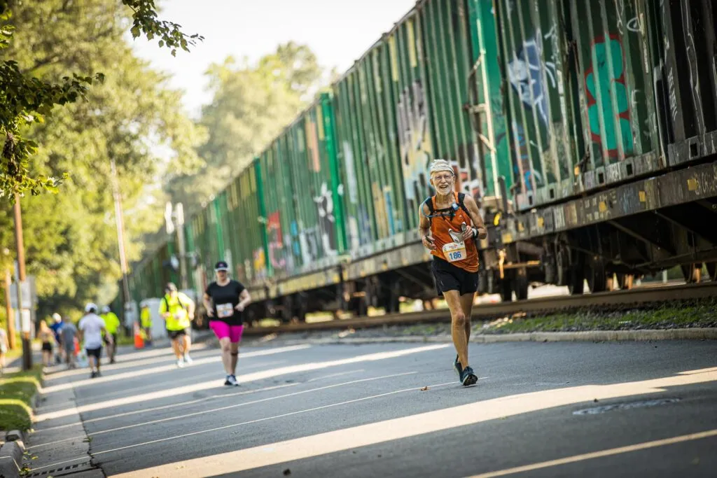 A male runner with a beard smiles as he runs past a long, graffiti-covered train on a sunny day.