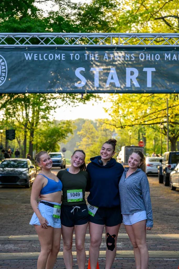 Four smiling women stand at the start line of the Athens Ohio Marathon, with a banner overhead that reads 'Welcome to the Athens Ohio Marathon START'.