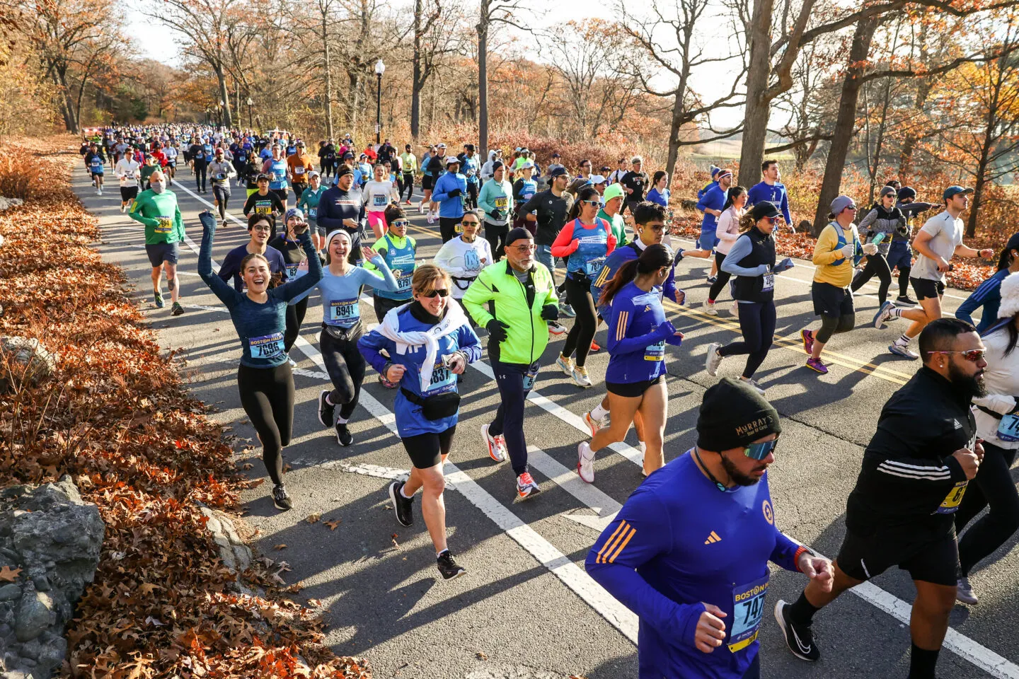 A large group of runners participating in a half marathon, with several runners in the foreground showing excitement and joy as they run along a tree-lined road.