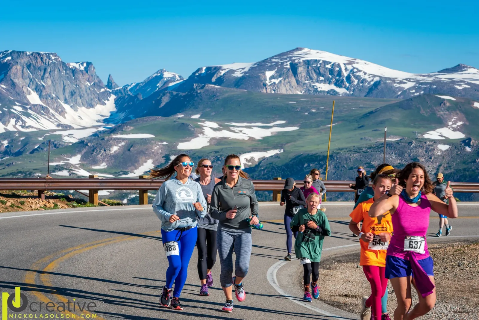 Runners smiling and giving thumbs up while racing on a scenic mountain road with snow-capped peaks in the background.