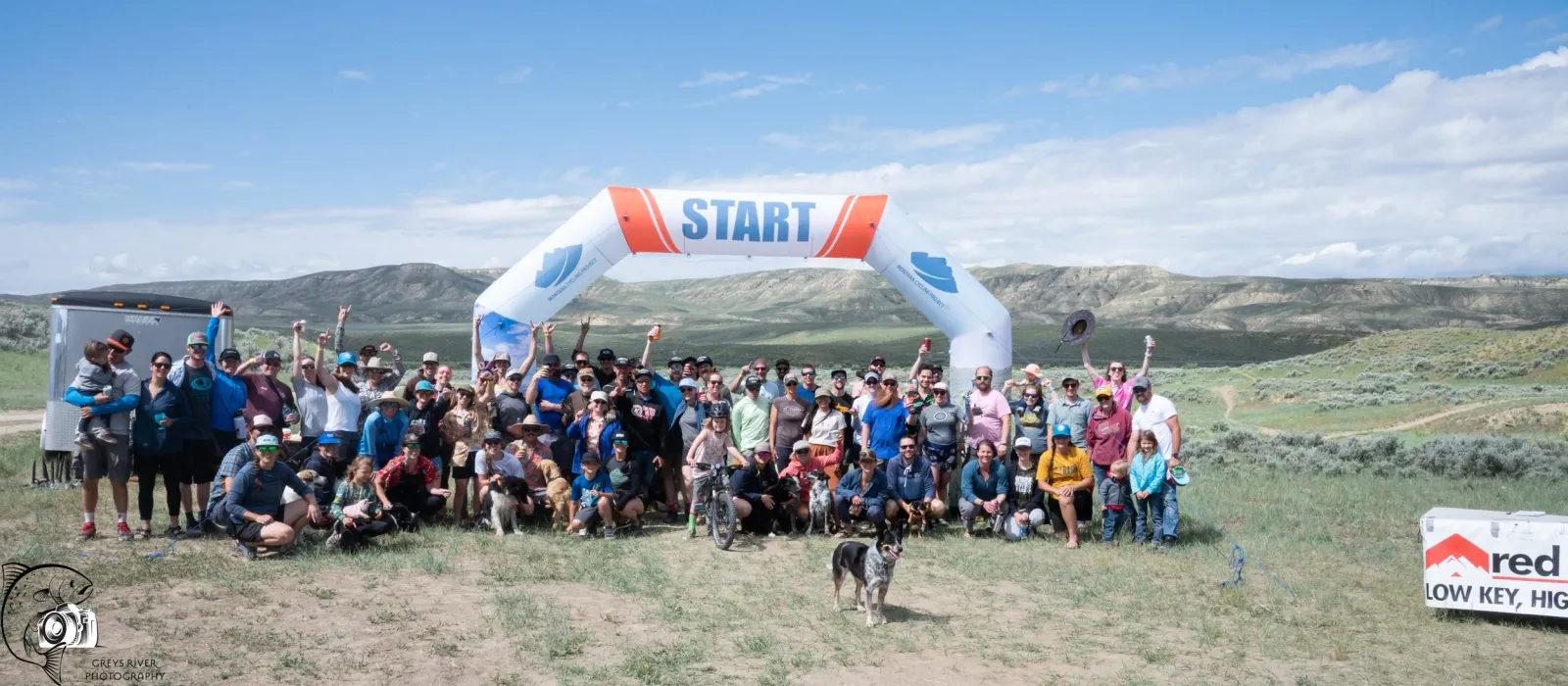 A large group of runners and spectators pose for a photo under a 'START' archway, with rolling hills in the background.
