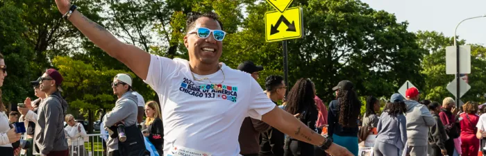 A smiling runner in a white t-shirt with sunglasses and arms raised in celebration, with a crowd of spectators in the background at the Bank of America Chicago 13.1 race.