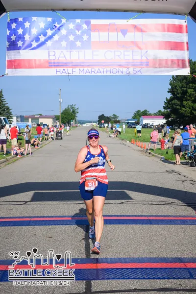 A female runner wearing a patriotic-themed outfit crosses the finish line at the Battle Creek Half Marathon & 5K, with spectators cheering on the sides.